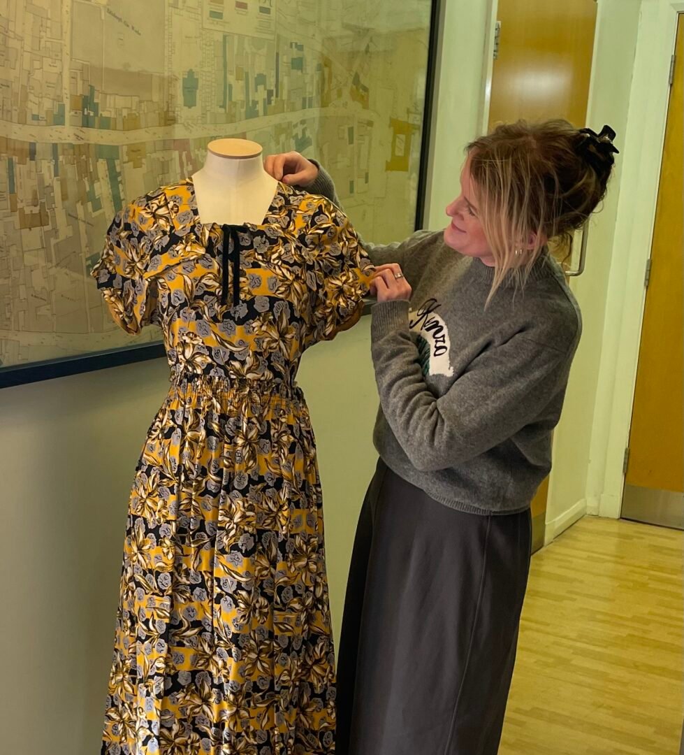 Vicky Garrington standing beside a tailor's dummy on which is displayed a black, grey and gold shortsleeved dress