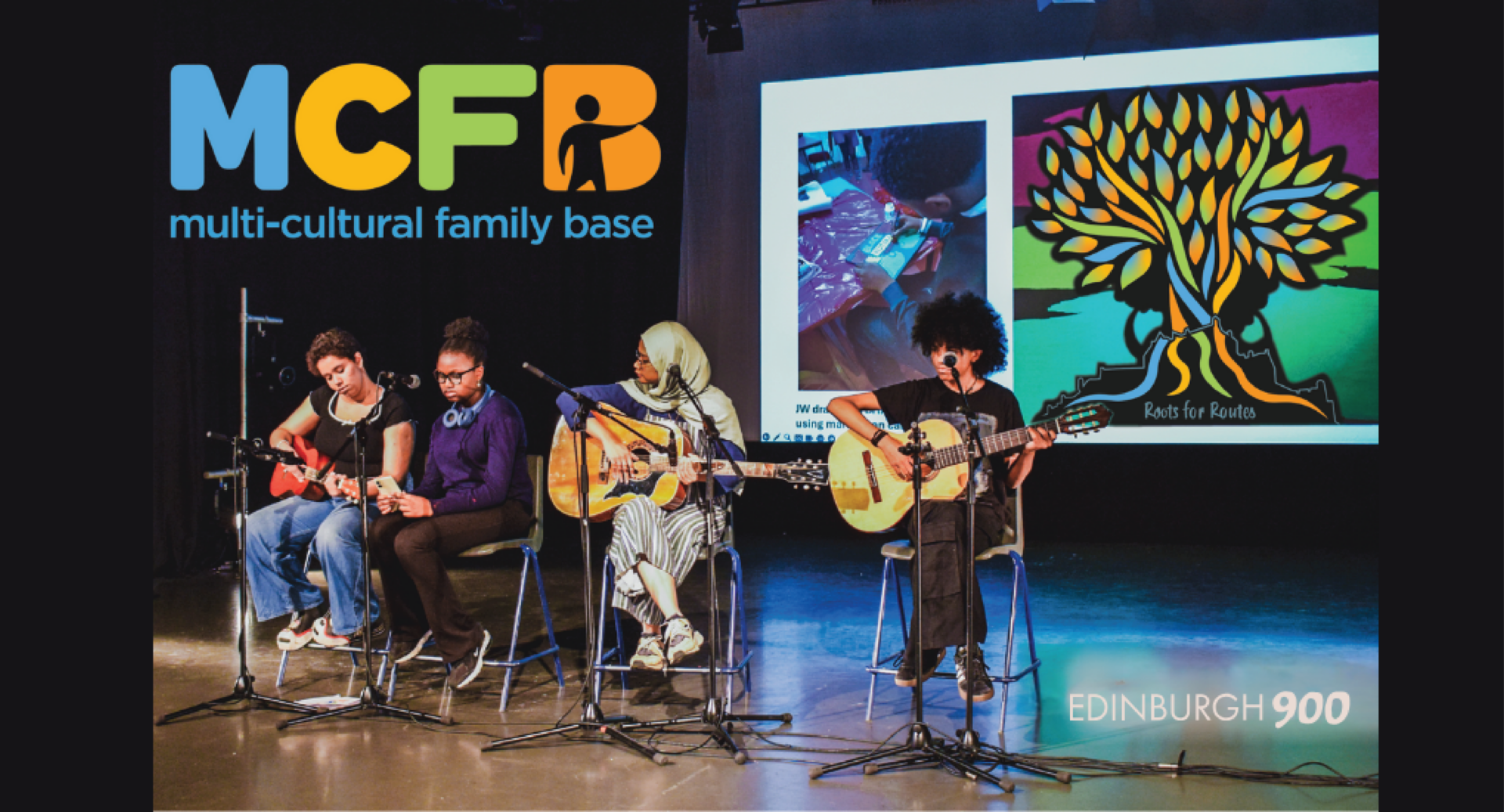 Four people sit on a stage, three with acoustic guitars, one holding a piece of paper. The backdrop is a photo of someone doing crafts and the Roots for Routes tree logo