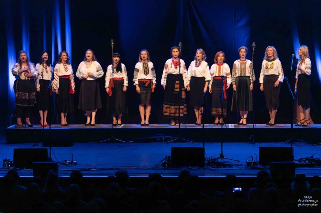 12 women on stage, all dressed in white shirt with embroidered detail and black skirt.