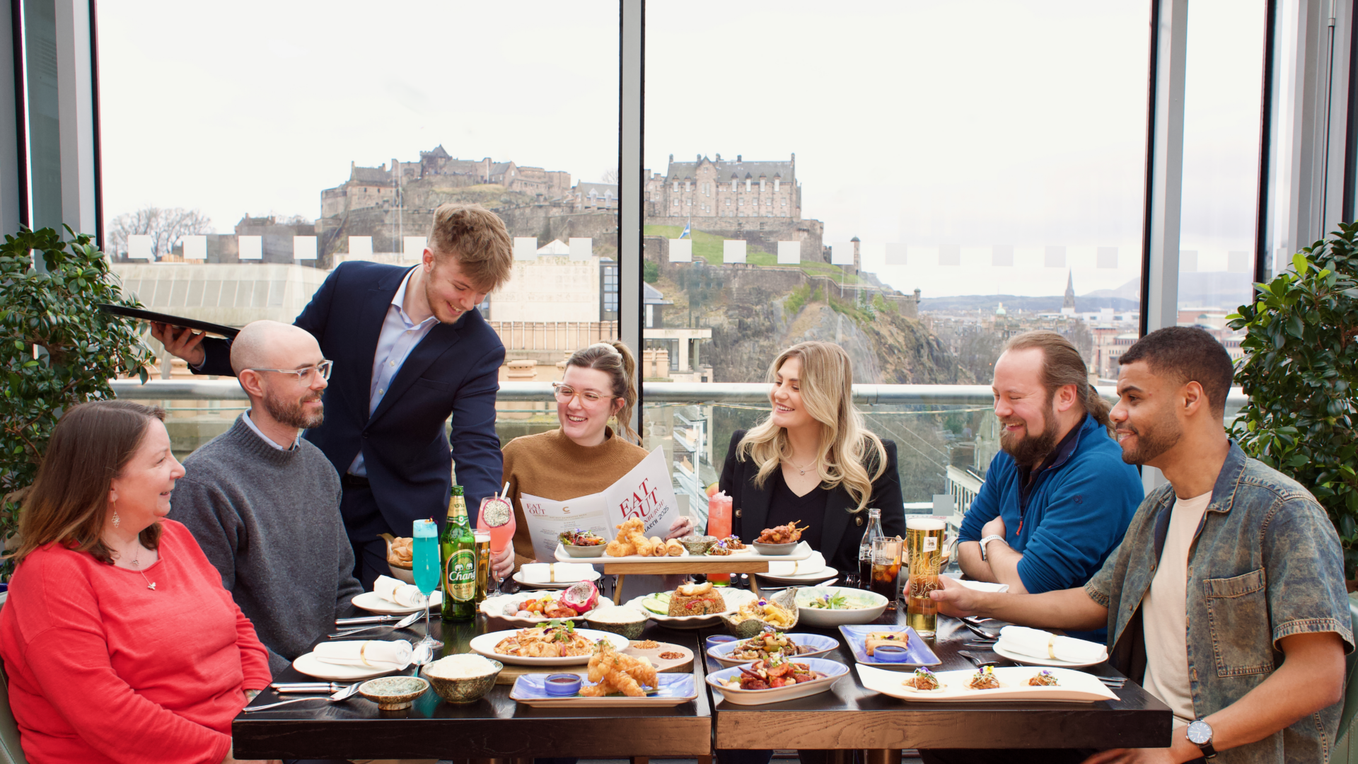 Group of 6 people sitting round a dinner table, with a waiter standing at the side. In the background is a view of a castle.