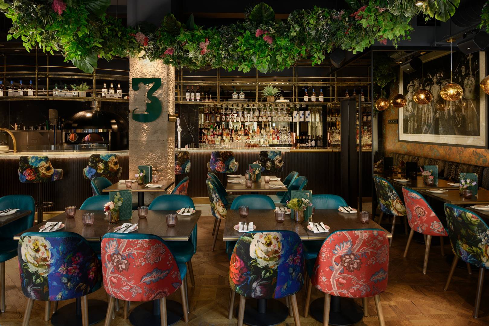 Luxury interior of a restaurant with multiple tables with bright, fabric covered chairs. There is foliage adorning the ceiling, a well stocked bar and warm lighting. ,© Photography by ZAC and ZAC