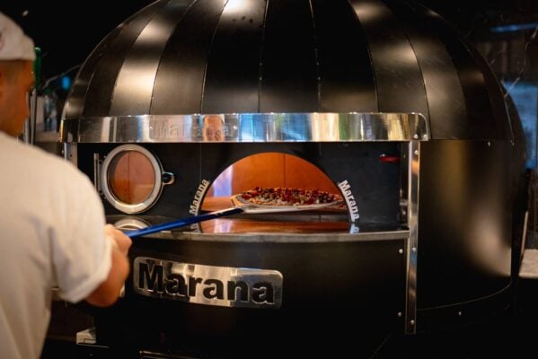 A pizza chef puts a pizza into a large professional pizza oven using a paddle.,© 3 Blind Mice