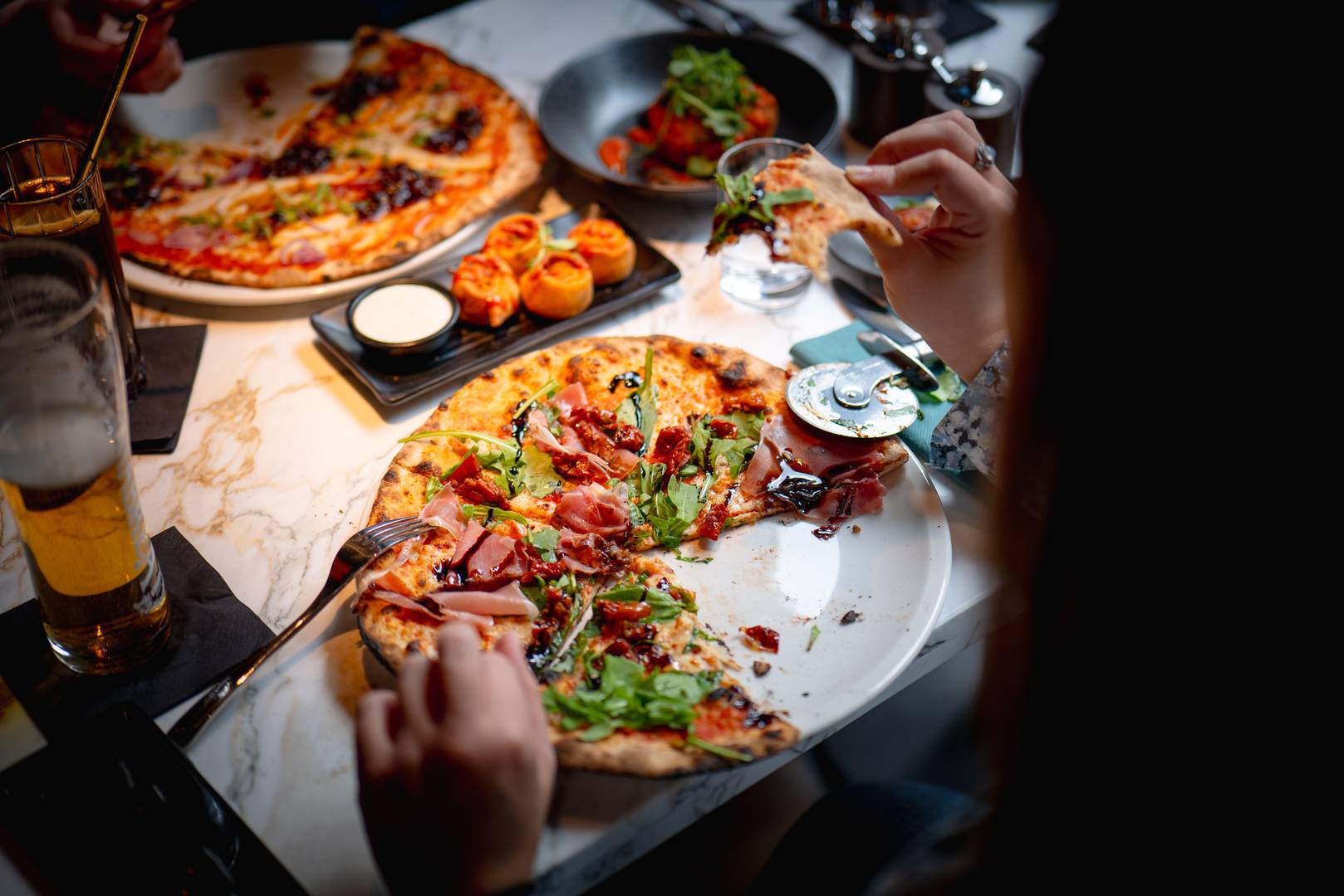 Someone eating a pizza topped with proscuitto. She holds a half-eaten slice of pizza and a pizza wheel can be seen on the table below. There is someone else dining opposite her. ,© 3 Blind Mice