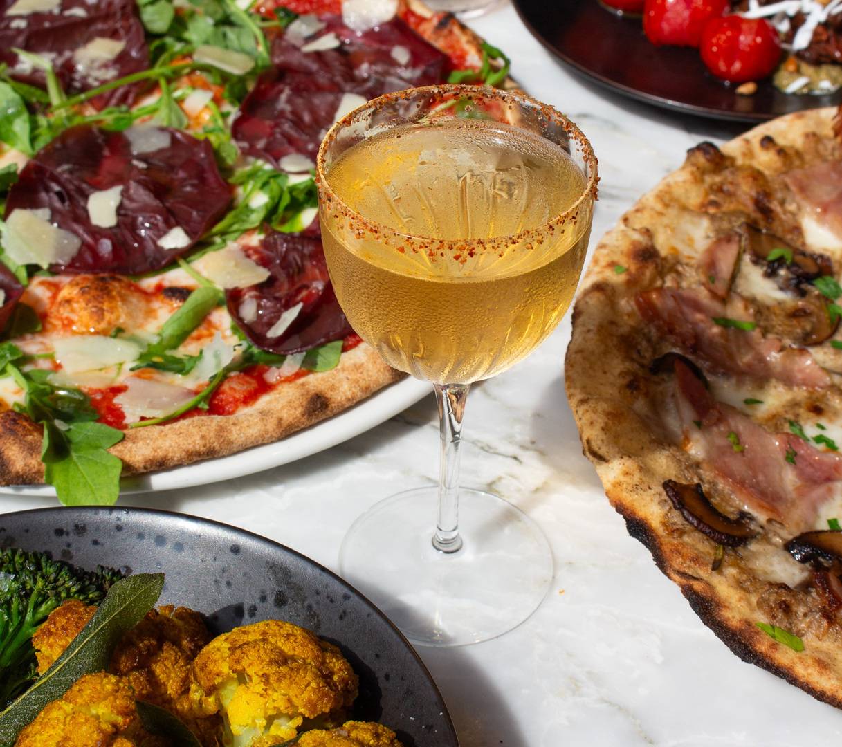 A spicy margarita in a cocktail glass, rimmed with tajín. In the background, there are various pizzas and small plates. ,© 3 Blind Mice