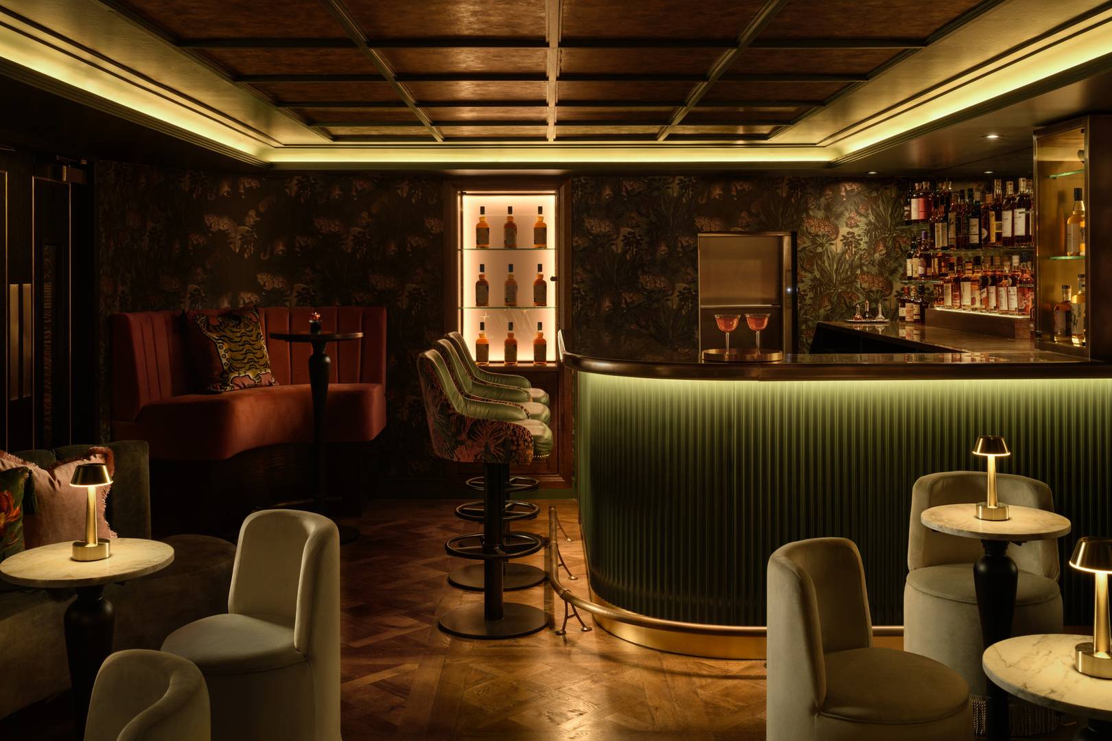 A warm, softly lit bar with floral-patterned wallpaper, a curved bar front clad in ribbed green paneling, and backlit shelves displaying bottles. Plush velvet chairs and couches in rich tones surround small round tables topped with brass lamps on a parquet wood floor.,© The Burrow - Photography by ZAC and ZAC