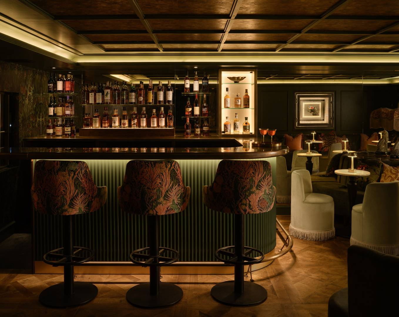 Softly lit bar with backlit shelves of whisky. There are textured upholstered bar stools in rich colours, and velvet seting around white curcular tables with brass lamps. ,© The Burrow - Photography by ZAC and ZAC