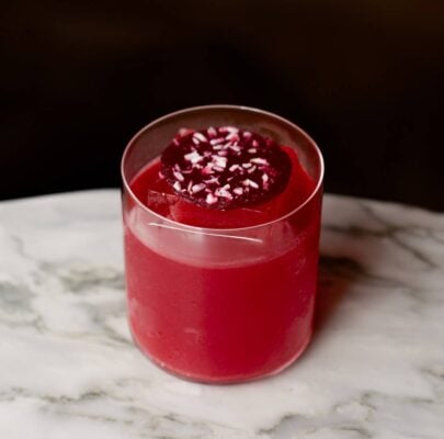 A pink cocktail with one large block ice cube, and a pink circular garnish with coconut sprinkled on top. It sits on a white marble table against a black background. ,© The Burrow