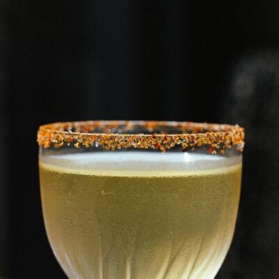 A close-up photo of a spicy margarita with a tajin rim. The cocktail is in a fancy cocktail glass against a dark background. ,© The Burrow