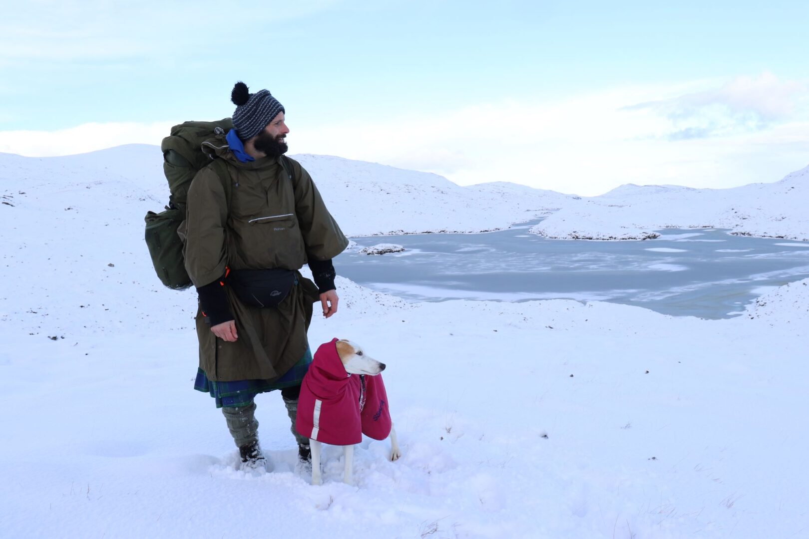 Man wearing winter clothing standing in a snowy landscape. At his feet is a small dog, wearing a red coat.