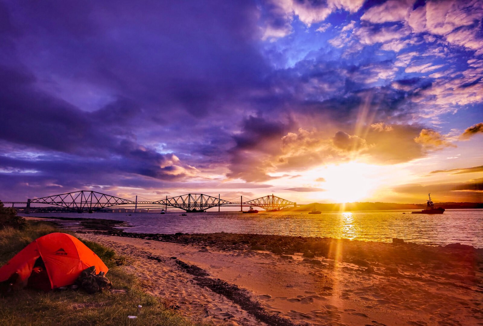 Sunset image of coast, with bridge in background and tent on the shore.