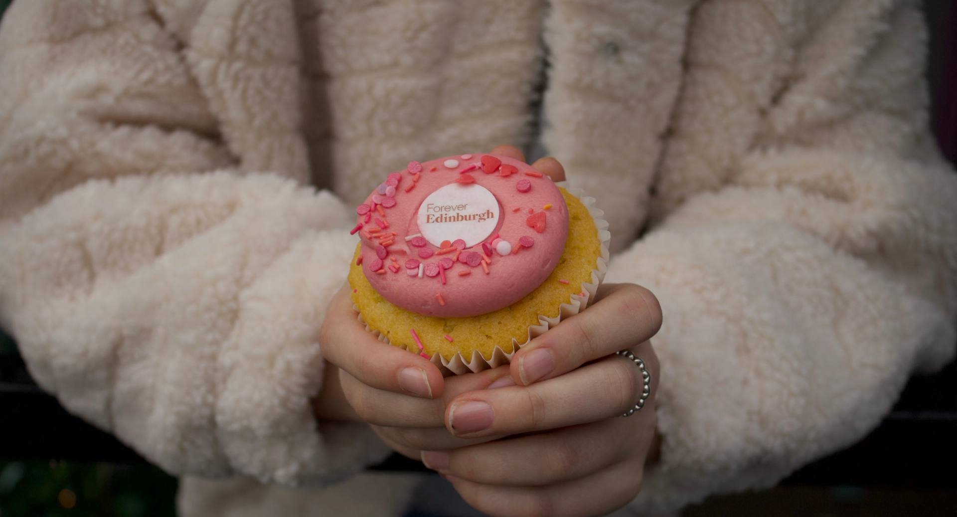 A female customer holds a Forever Edinburgh branded Mimi's Bakehouse cupcake with pink icing and the Forever Edinburgh logo.