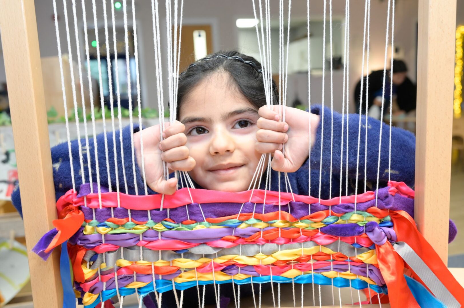 Young girl looking through a loom, which has brightly coloured ribbons thread though it.