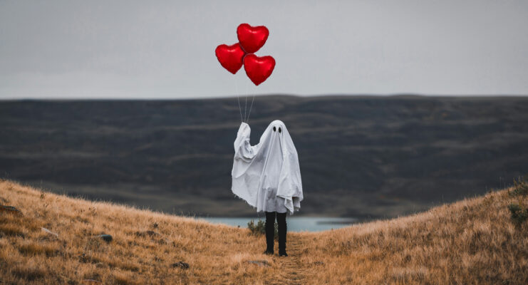 Person dressed with a sheet over them to look like a ghost carrying red heart balloons on a hill with view to reservoir