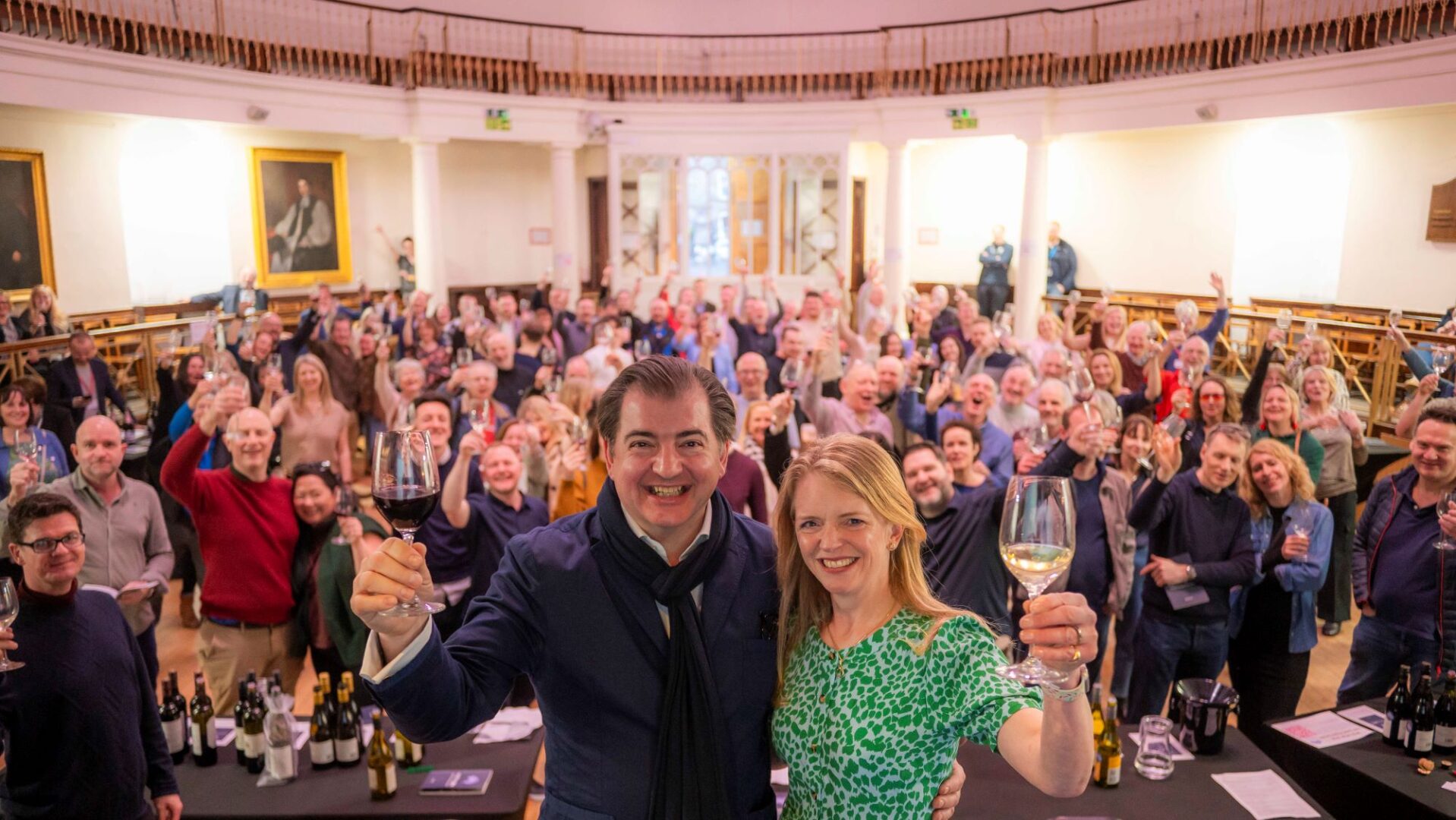 Man and woman standing in front of a room of crowded people, all raising wine glasses.