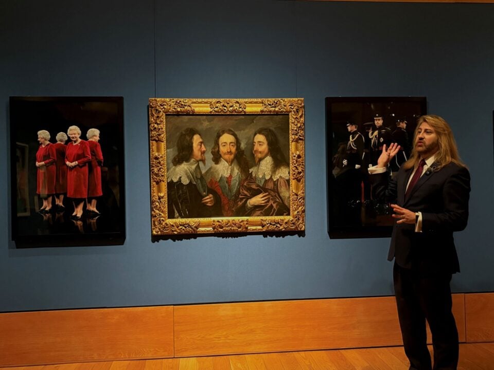 Man standing against a wall, on which hang 3 portraits.