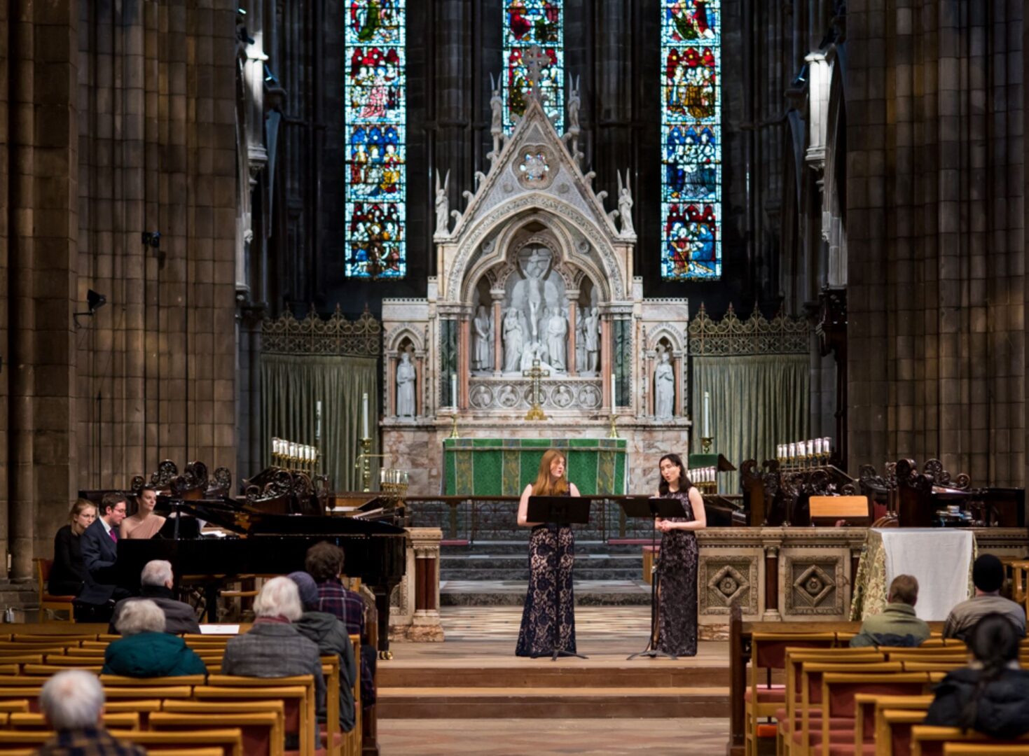 In interior of a cathedral, with stained glass window and alter. 2 women are standing at lecterns in front of a small crowd of people.