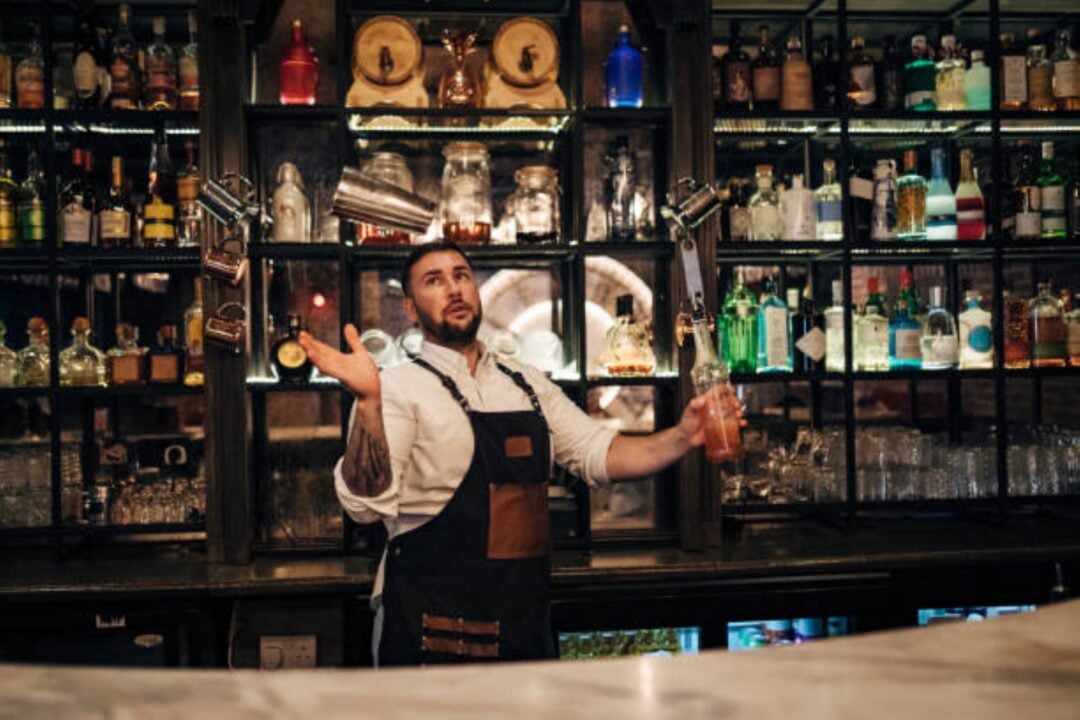 Man standing at a bar throwing a cocktail shaker in the air. Behind him is a wall filled with glass bottles.