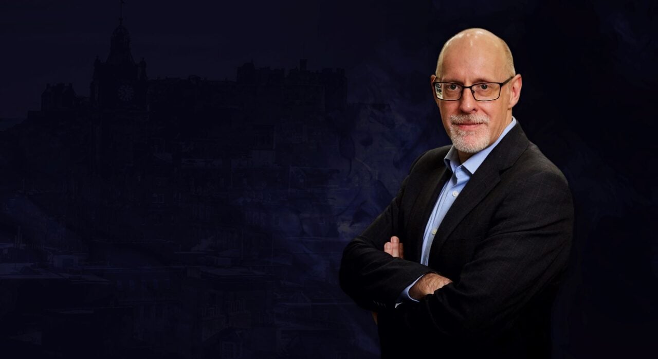 Man wearing dark suit and pale blue shirt standing with arms folded, against a background of a dark city sky outline.