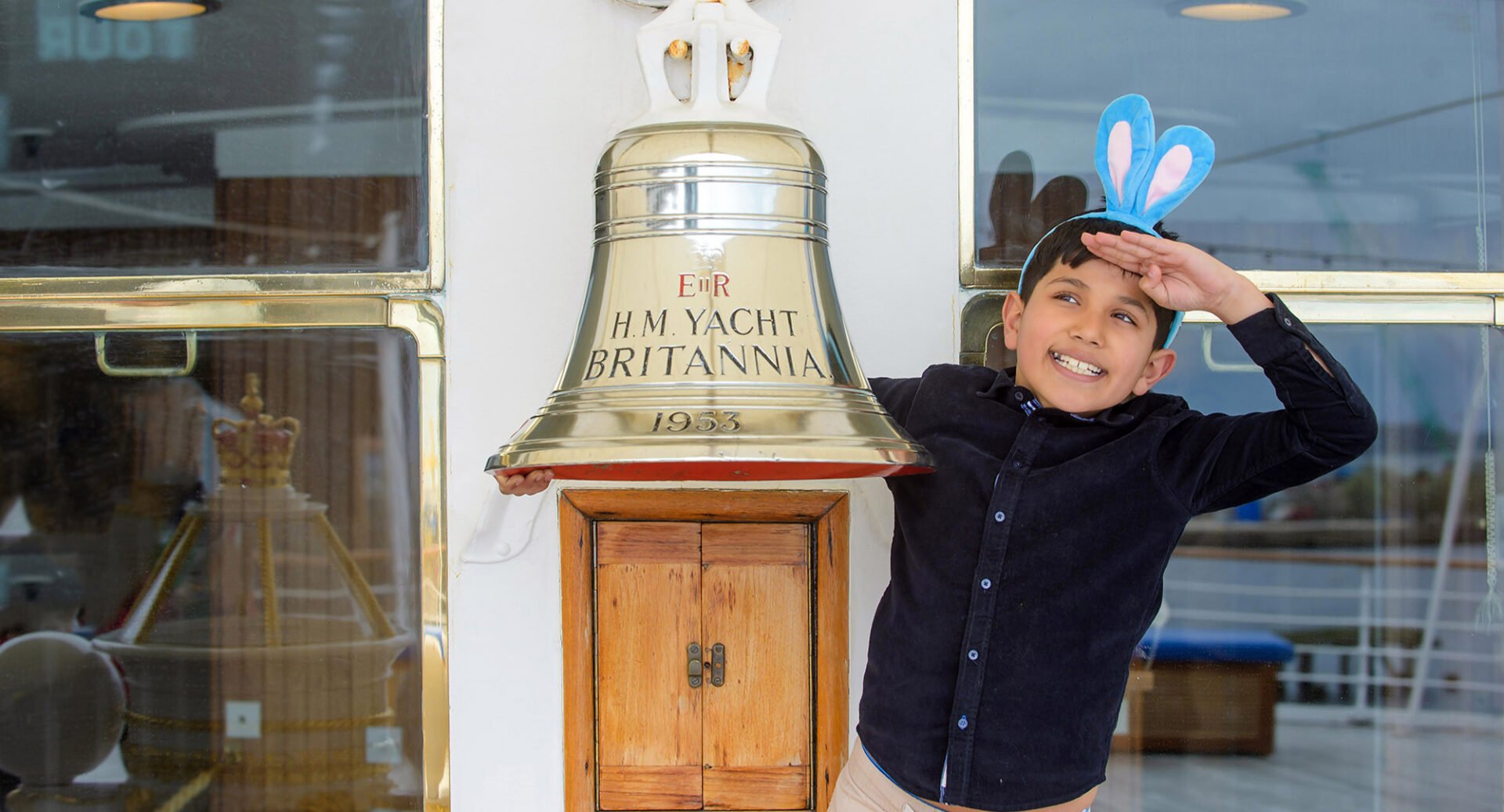 Yong boy wearing bunny ears standing beside a large brass bell, onboard a boat.