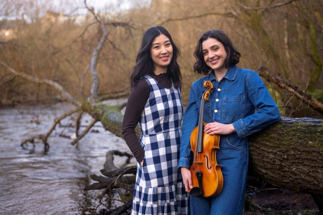 2 women, one holding a viola, standing at the side of a river.