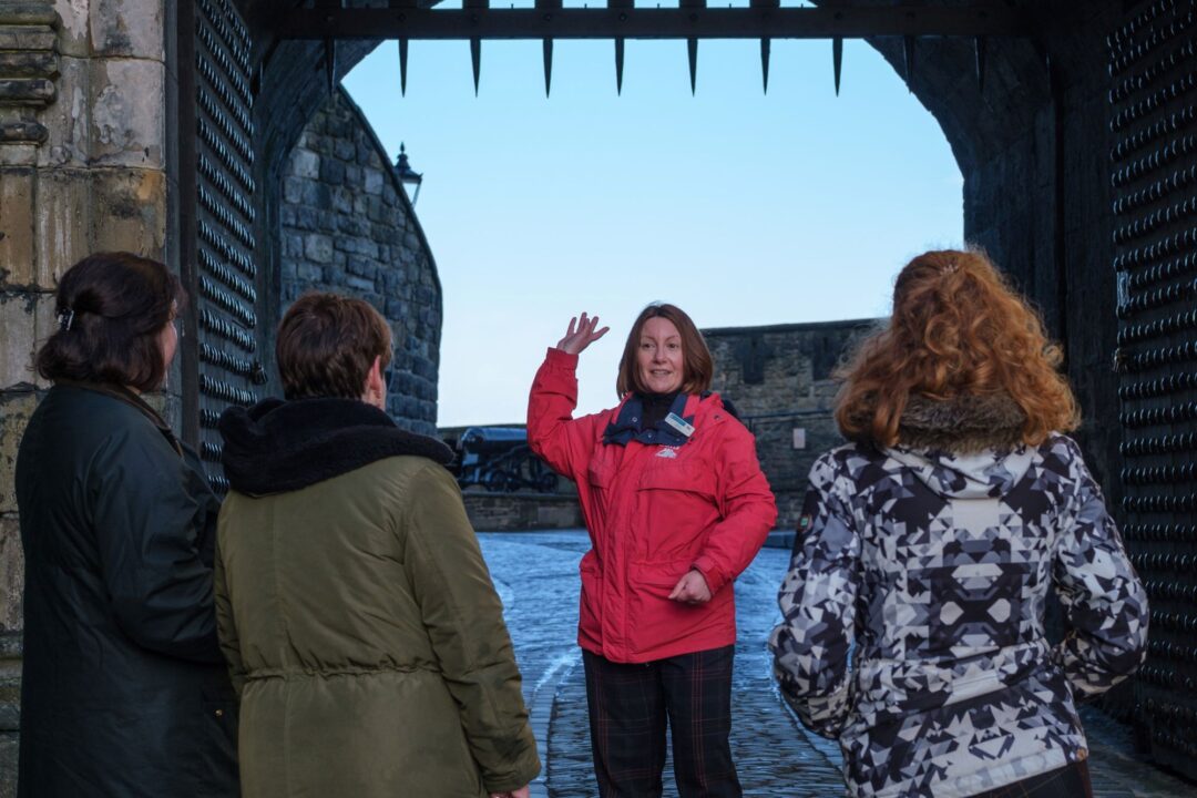 Women in a red jacket standing in front of a group of 3 people, pointing to something behind her.