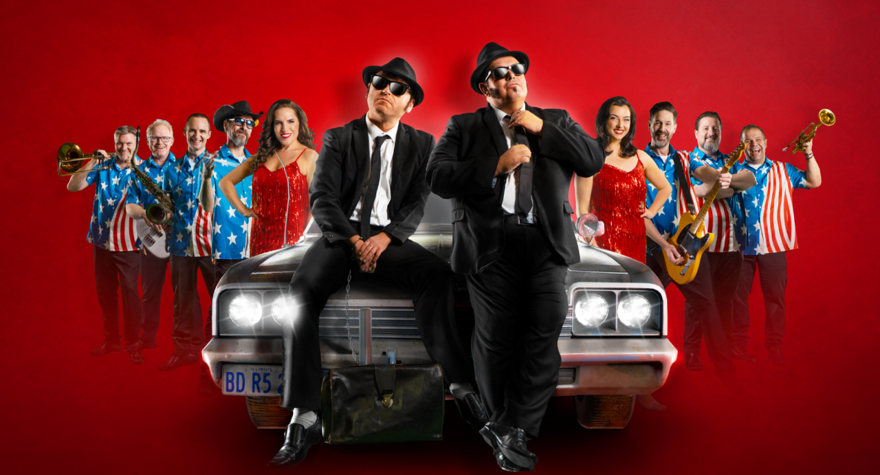2 men dressed in black suits, hats and sunglasses, standing in front of a car. Of either side of them are musicians.