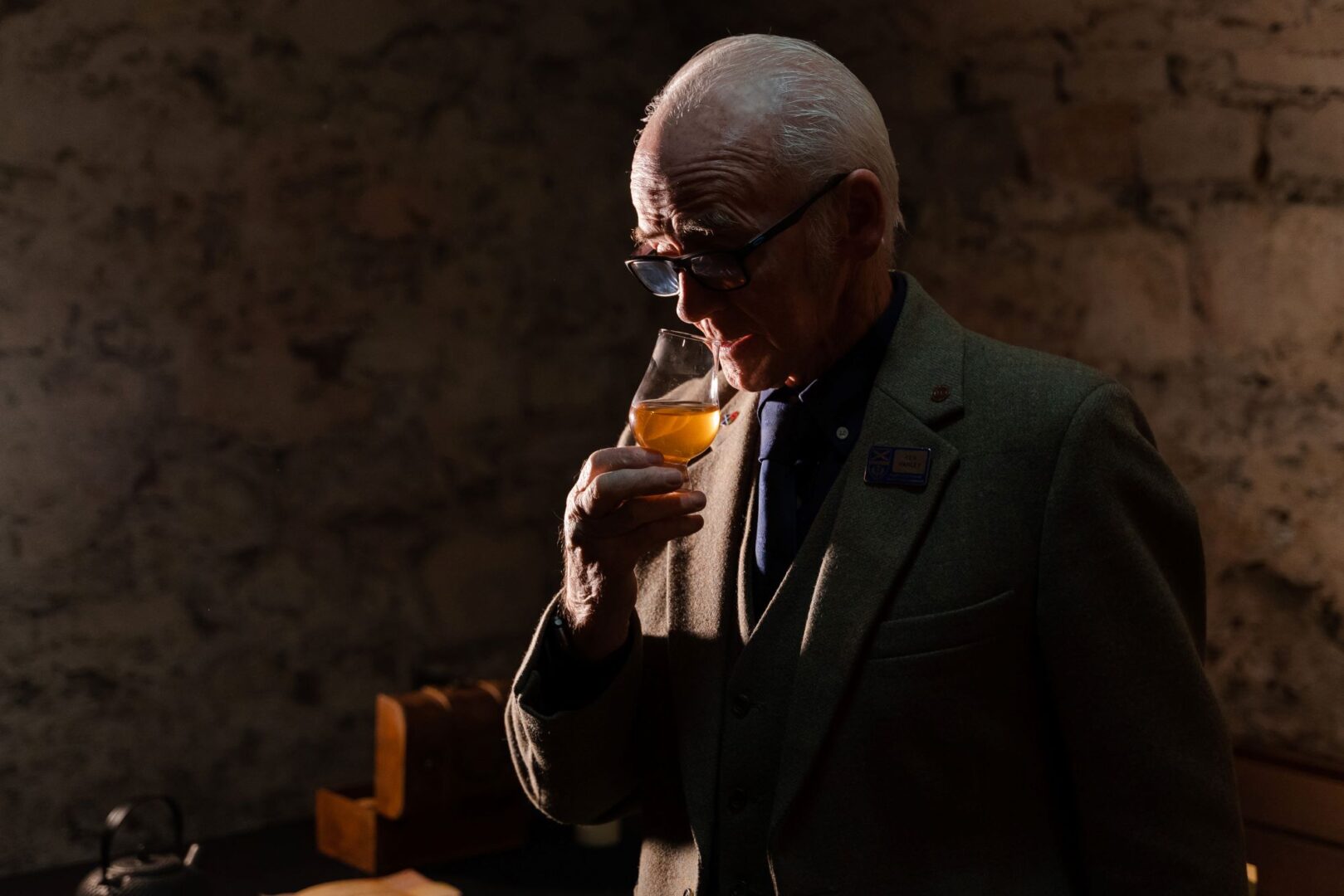 A man holding a small glass filled with amber liquid and raising it to his nose.
