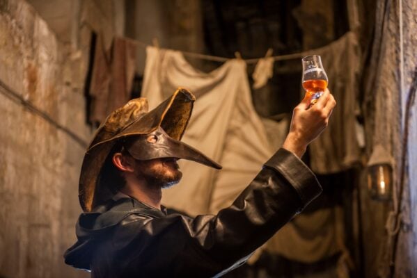 Person wearing a mask with a pointy nose holding aloft a small glass filled with amber liquid.