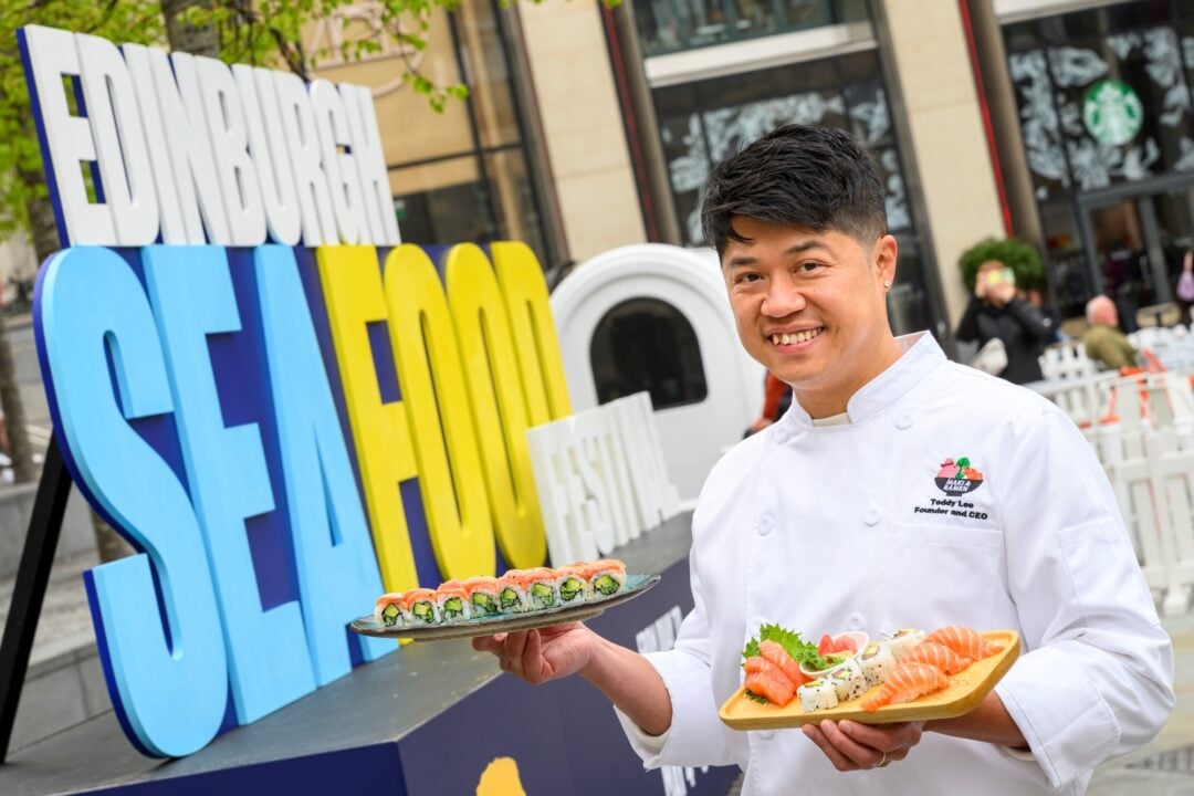 Chef holding 2 plates of sushi, standing in front of a large sign saying Edinburgh Seafood Festival.
