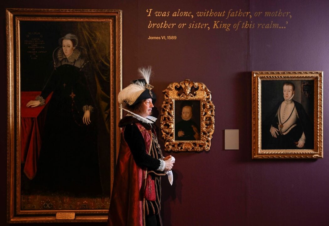 Man wearing a long elaborate clock and feathered hat walking past a wall, on which a collection of portraits.