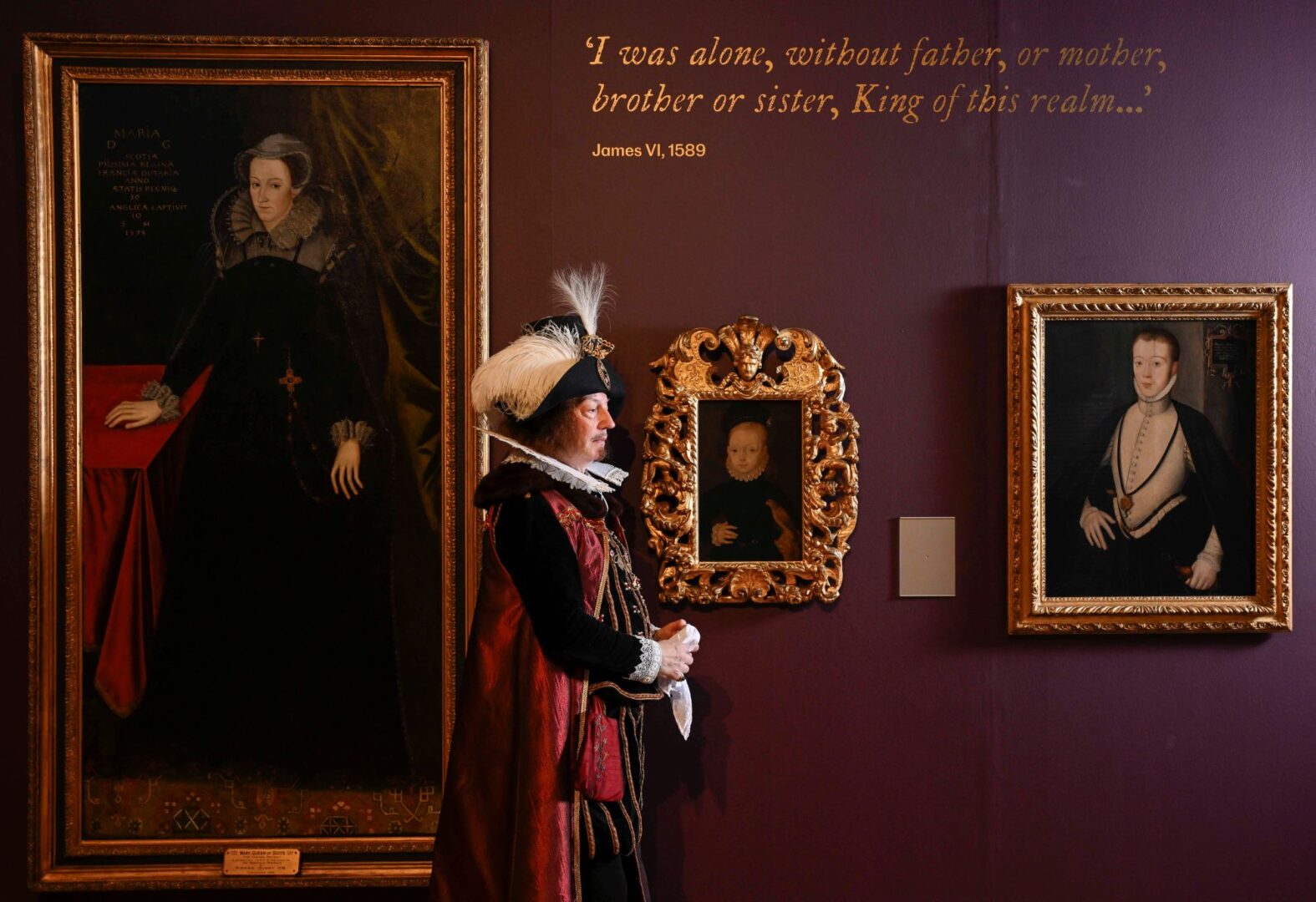 Man wearing a long elaborate clock and feathered hat walking past a wall, on which a collection of portraits.