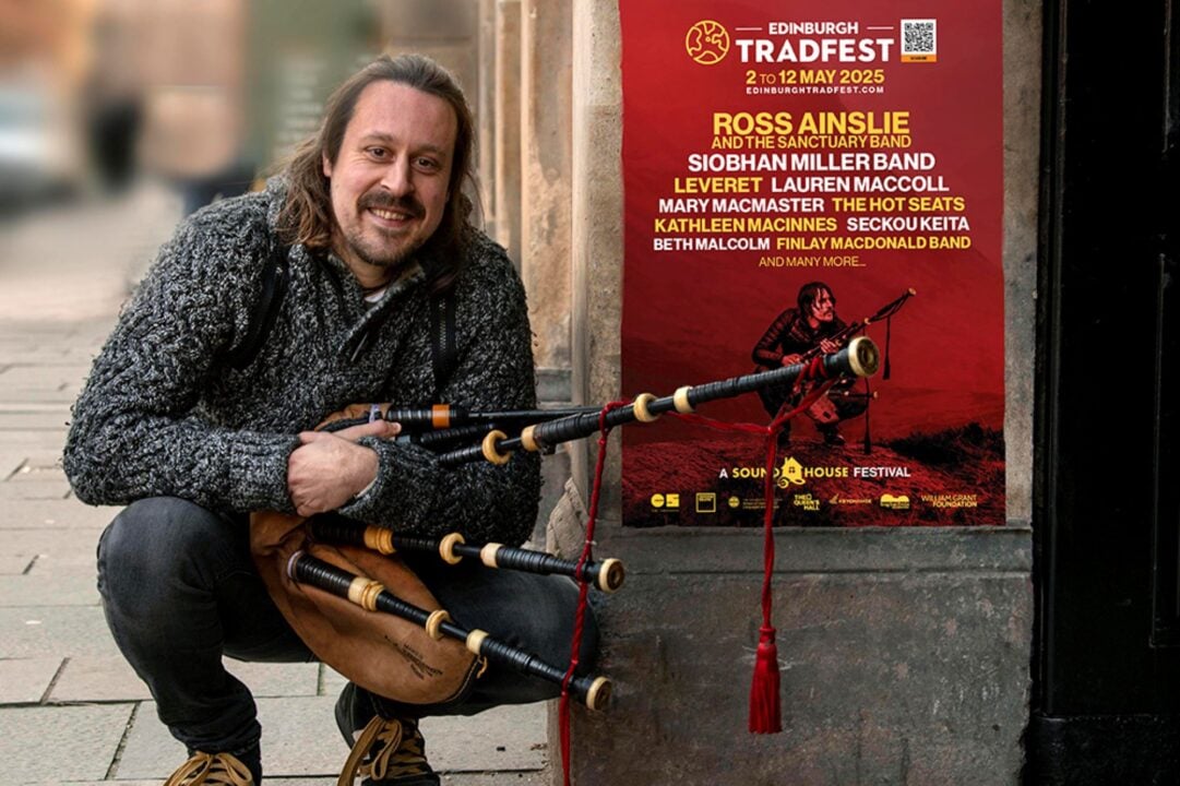 Man holding a set of bagpipes, crouching down beside a stone wall.