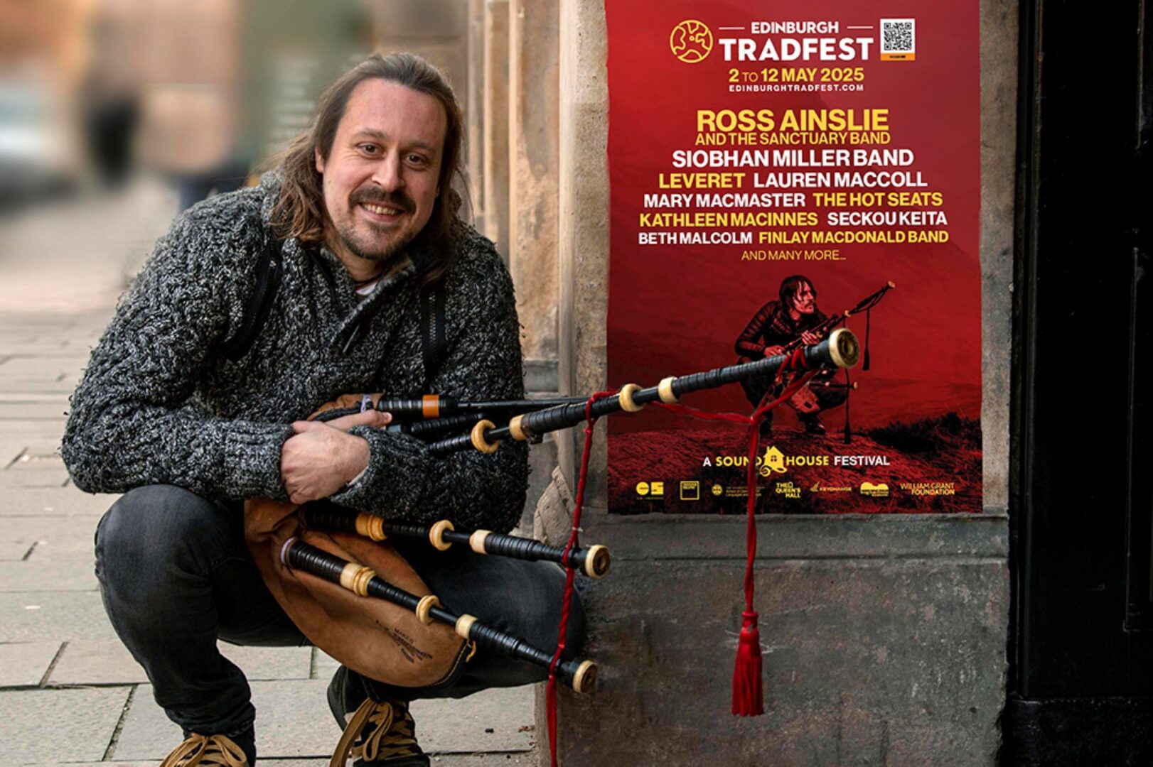 Man holding a set of bagpipes, crouching down beside a stone wall.