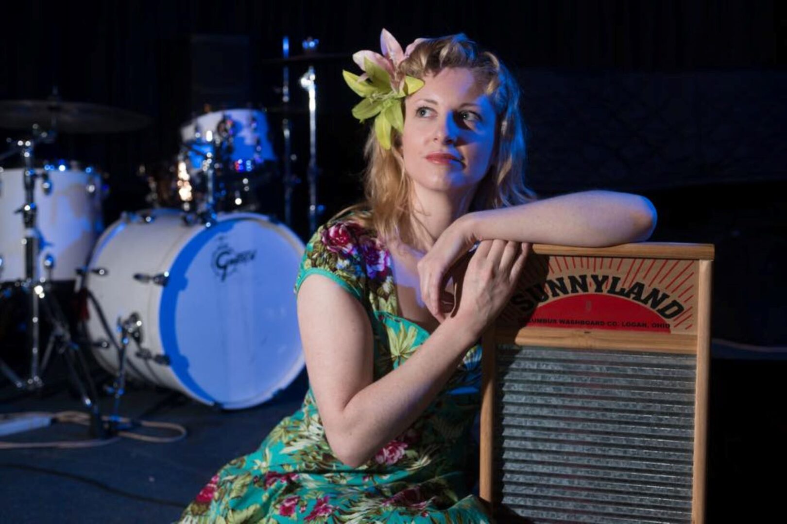 Women wearing a colourful dress and flower in her hair, sitting down, with a set of drums in the background.