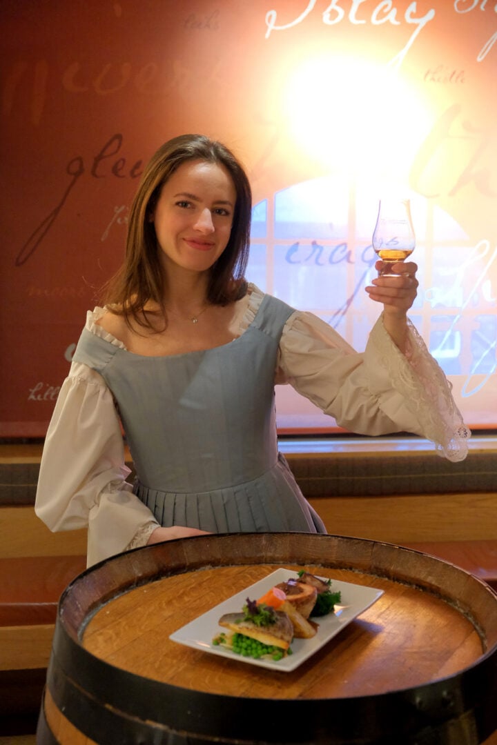 Woman wearing an old-fashioned blue pinafore with white blouse, standing in front of a barrel, on which is a plate of food. She is also holding a glass in one hand.