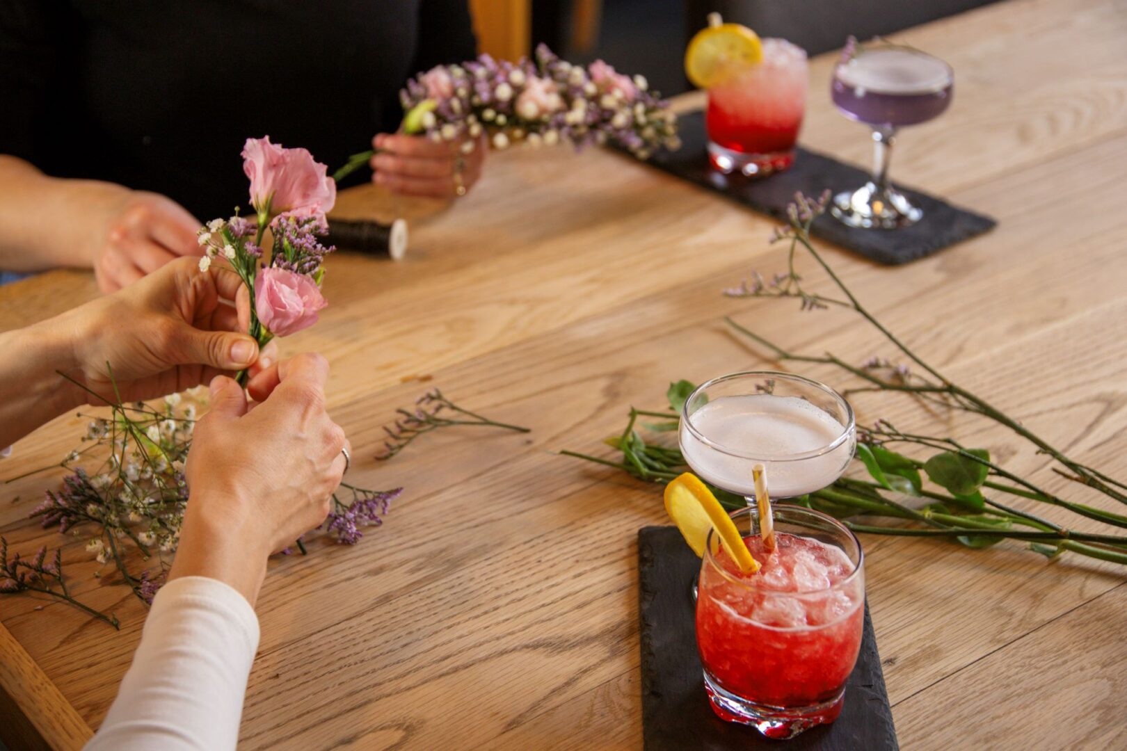 A pair of hands showing making a flower garland. On the table are also glasses with brightly coloured drinks.