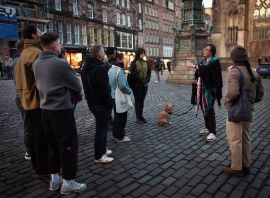 The tour guide talking to a crowd of people. He is mid-story outside St. Giles Cathedral, a stop on the free walking tour and the LGBTQ walking tour. The guide is holding a pink umbrella and a dog is present