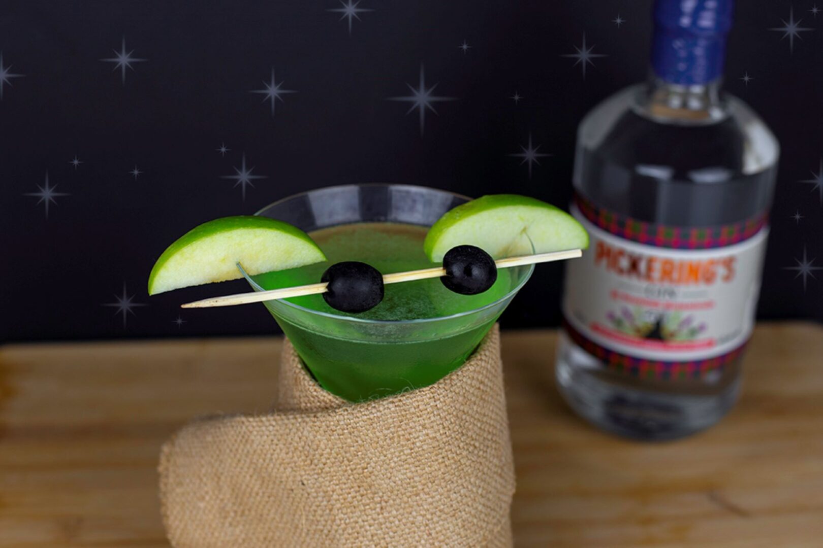 Green coloured drink in glass, garnished with 2 black olives and slices of green apple. In the background is a bottle of gin.