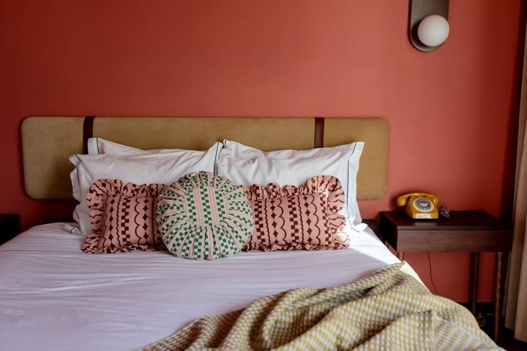 This image is showing a bed with an olive suede headboard against an apricot coloured wall. The bed has white linen bedding with 3 TBCo cushions, two are pink rectangles with dark pink squiggles and shapes, the third cushion is circular with light pink stripes and green squares. ,© tbco