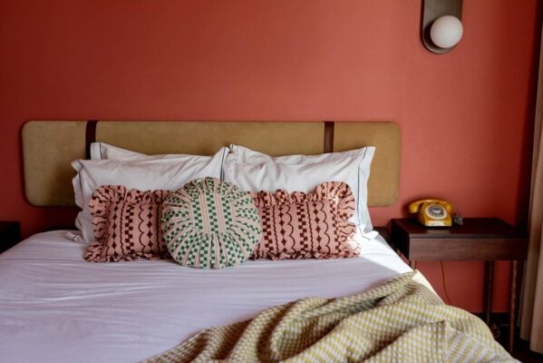 This image is showing a bed with an olive suede headboard against an apricot coloured wall. The bed has white linen bedding with 3 TBCo cushions, two are pink rectangles with dark pink squiggles and shapes, the third cushion is circular with light pink stripes and green squares. ,© tbco