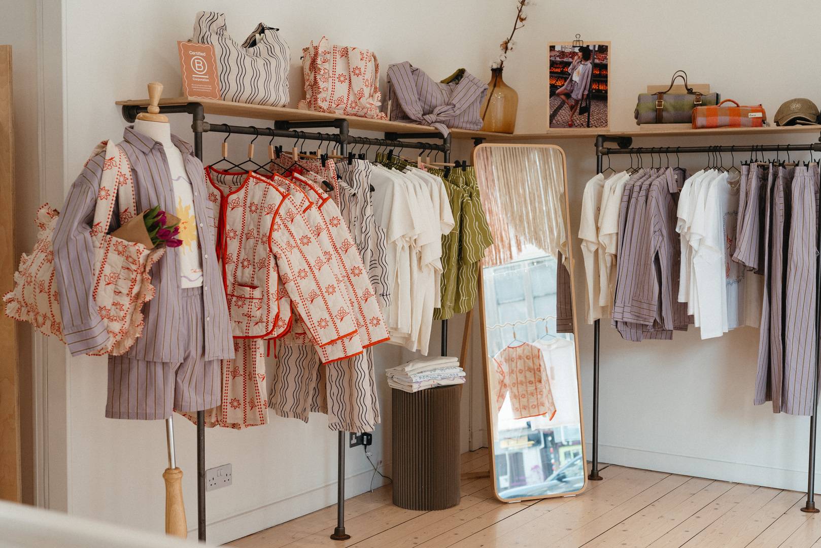 This image shows inside the tbco store, the top shelf has 3 different coloured cotton bags on display. Hanging up there are lilac striped shirts and trousers, and an assortment of white and cream T-shirts. ,© tbco