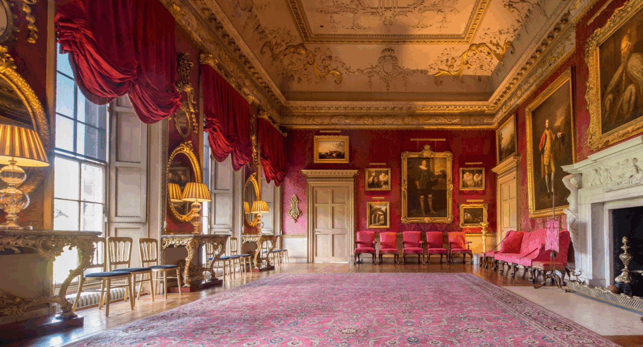 Grand, large room, with dark red walls, an ornate ceiling and large fireplace. On the walls are a series of paintings in gilt frames.