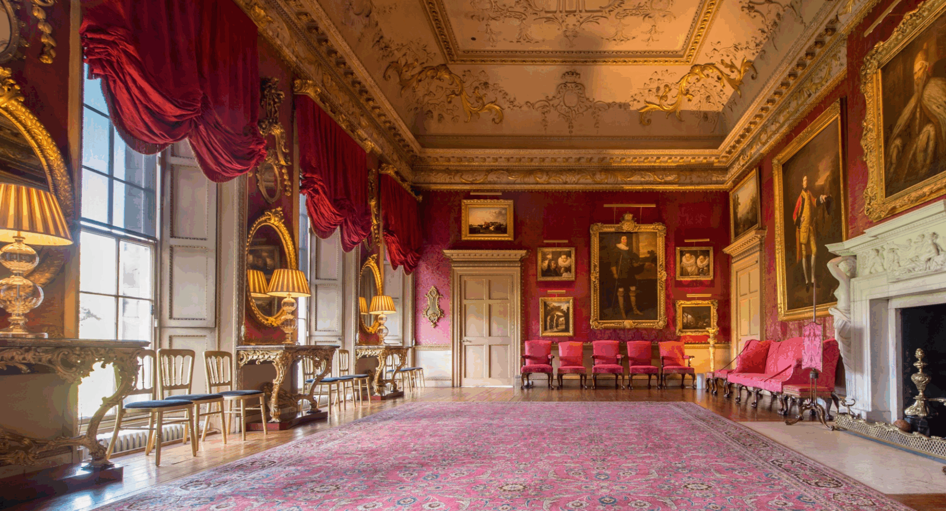 Grand, large room, with dark red walls, an ornate ceiling and large fireplace. On the walls are a series of paintings in gilt frames.