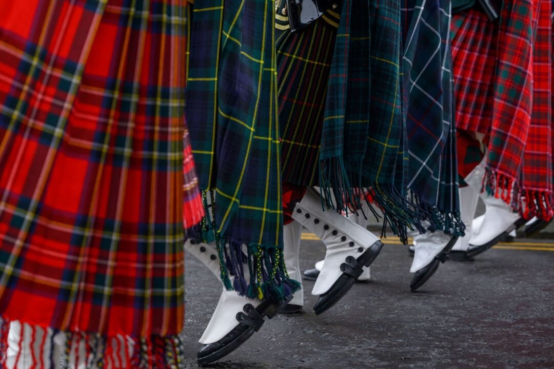 Ground level view of row of feet, wearing white and black boots, and long sashes of brightly coloured Scottish tartan.
