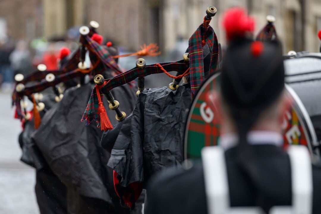 Back view of people marching, while playing bagpipes and playing drums, all wearing traditional Scottish dress.