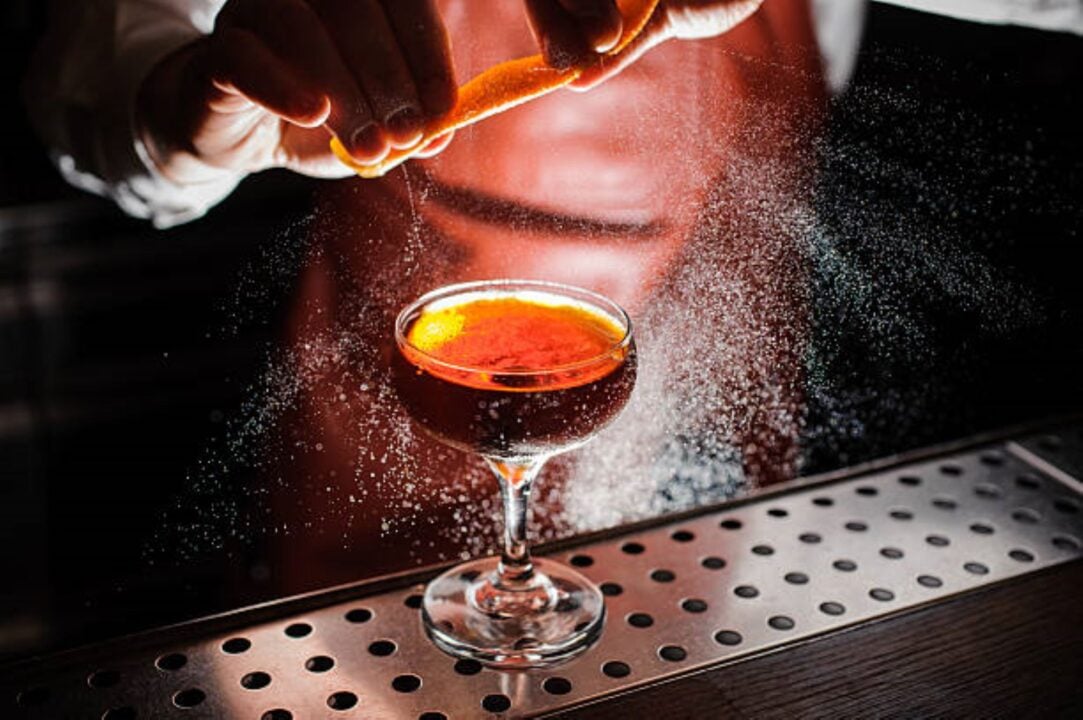 A figure standing over a steel counter, on which is a filled cocktail glass. The figure is squeezing a strip of orange rind over the drink.
