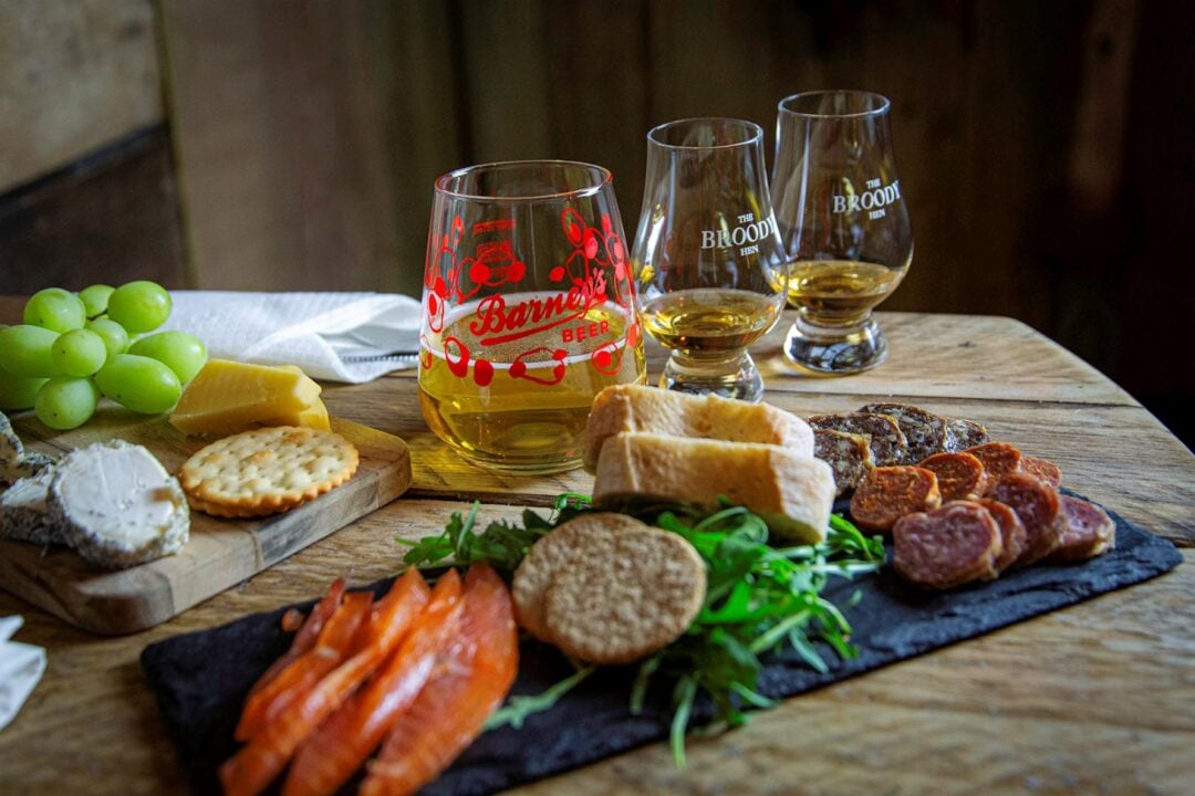 A table set with 3 glasses, filled with amber-coloured liquid, grapes, crackers and cheeses.