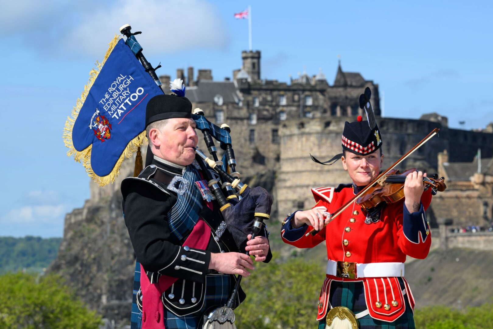 2 figures - one playing bagpipes, and one playing a violin, in traditional Scottish dress. Behind them is a large castle.