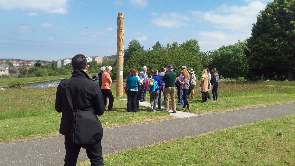 A group of people standing on a grassy area near a tall, carved sculpture with trees in the background