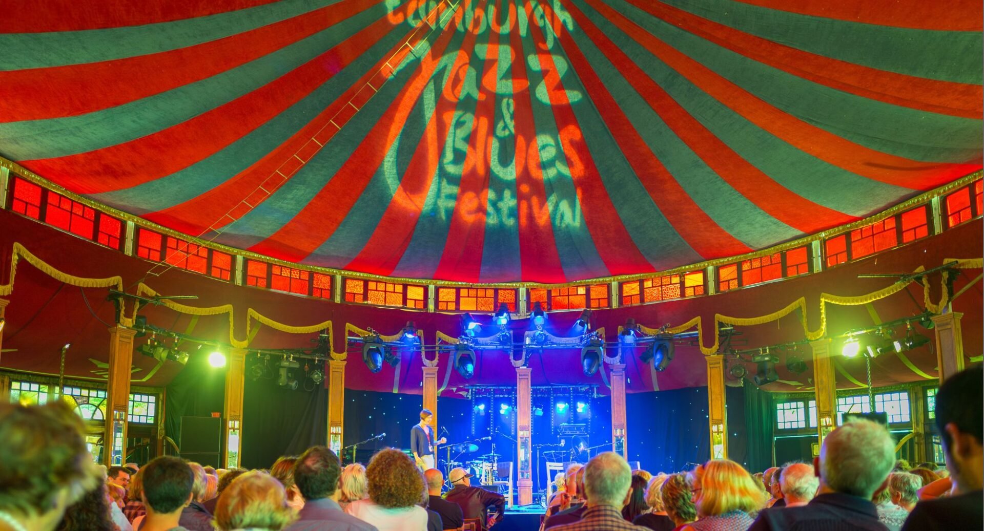 Group of people gathered in a large tent, withe large red and green stripy roof, watching musicians on the stage in front.